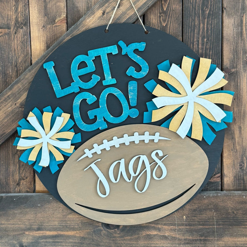 A round hanging wooden sign with a dark charcoal background, turquoise and light blue 'Let's Go!' lettering, a tan football shape with 'Jags' in white script, and two blue-and-tan pom-pom flowers, displayed against a rustic wood plank wall.