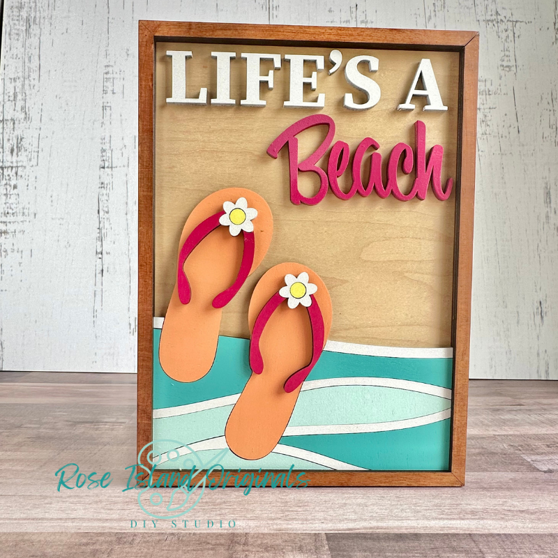 A framed DIY craft sign with a wooden frame and layered paper design that reads 'LIFE'S A Beach' in white and pink. Two orange flip-flops with small white daisy accents are layered over a tan and turquoise beach wave background.