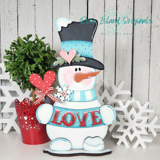Decorative snowman with a top hat, heart, and 'LOVE' sign next to a potted plant and snowflakes on a wooden background.