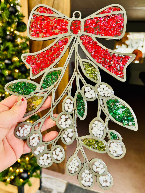 Decorative mistletoe ornament with red, green, and white beads held by a hand against a blurred Christmas tree background.