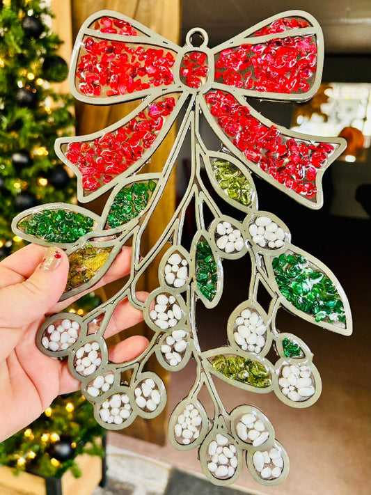 Decorative mistletoe ornament with red, green, and white beads held by a hand against a blurred Christmas tree background.