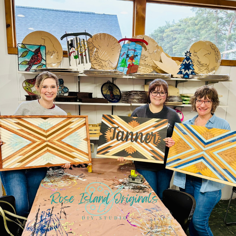Three women holding wooden art pieces in a workshop setting with 'Rose Island Originals' branding.