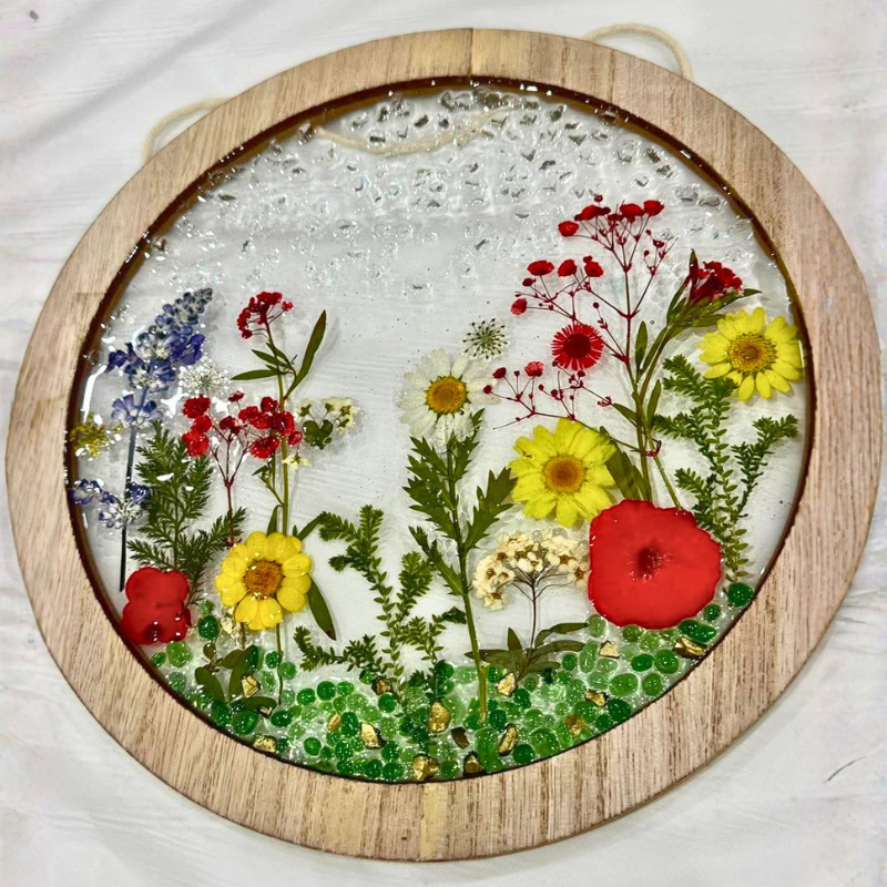A circular wooden-framed pressed flower wall hanging with preserved botanicals including yellow daisies, red poppies, blue flowers, green foliage, and clear resin droplets on a white background.