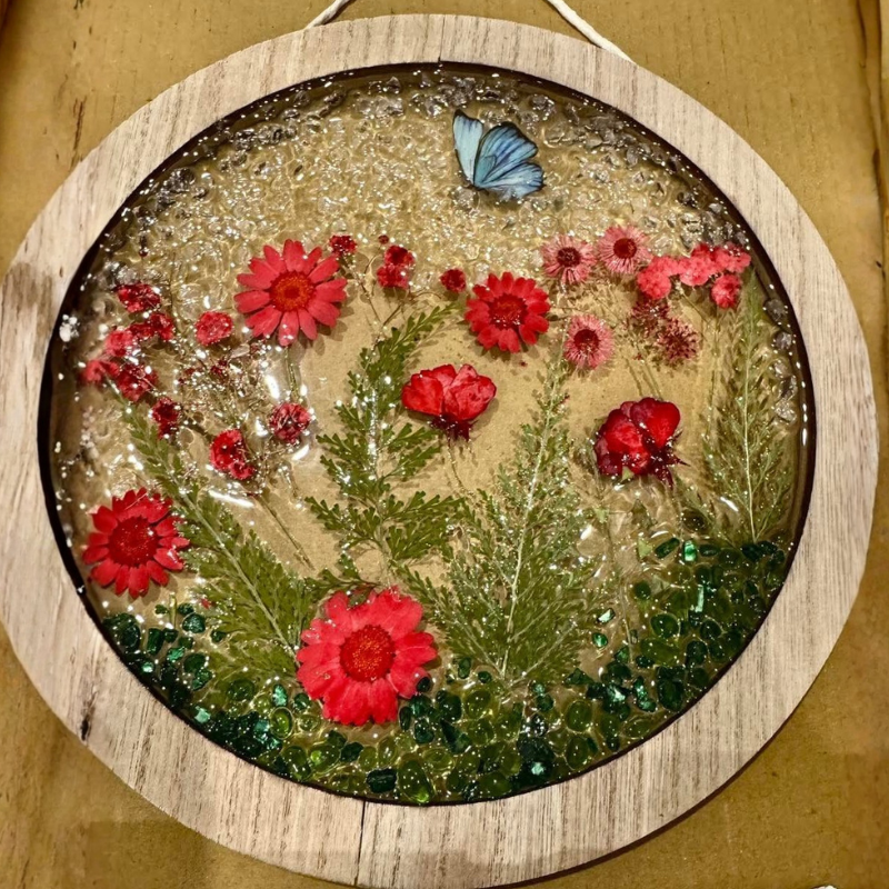 A round wooden hanging plaque featuring a pressed floral arrangement with red flowers, green foliage, glitter, and a small blue butterfly on a tan background.