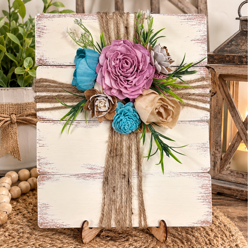 A decorative faux floral cross hanging on a distressed white wooden sign, styled with burlap ribbon, green leaves, and paper flowers in pink, blue, and beige, displayed on a rustic wooden surface with a lantern and plant in the background.