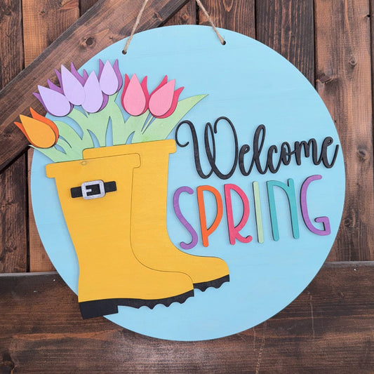 Decorative sign with yellow rain boots, tulips, and 'Welcome Spring' text on a wooden background.