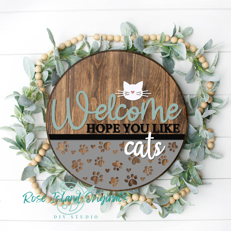 Decorative sign with 'Welcome Hope You Like Cats' text surrounded by greenery and wooden beads on a white background.