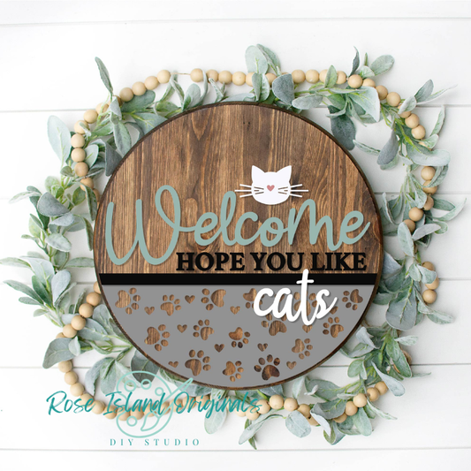 Decorative sign with 'Welcome Hope You Like Cats' text surrounded by greenery and wooden beads on a white background.