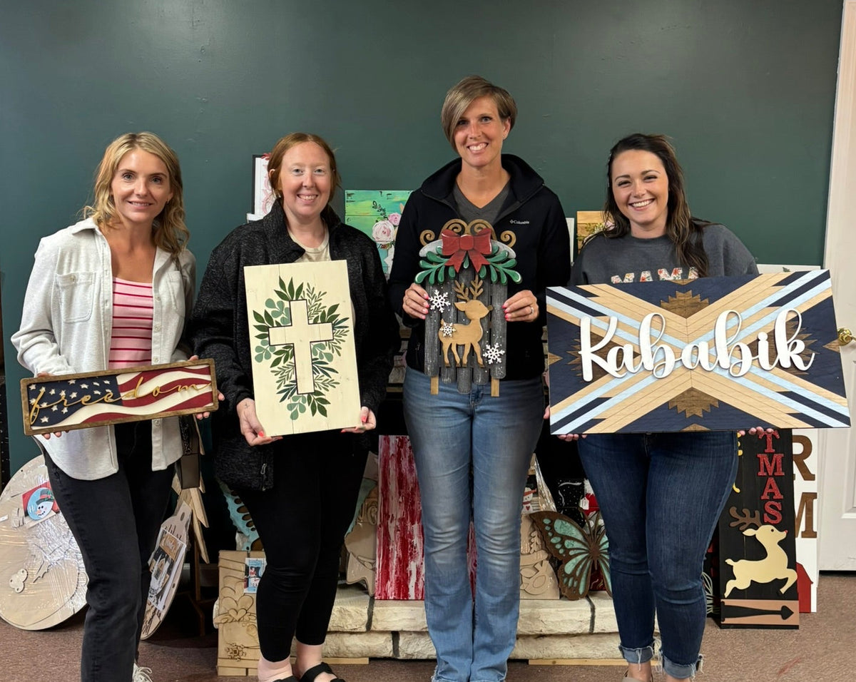 Four women standing together in a room, each holding a decorative sign or artwork. The signs include a rustic American flag, a botanical design, a colorful floral and deer motif, and a blue-and-white cross design with the word 'Rabikab' in script.