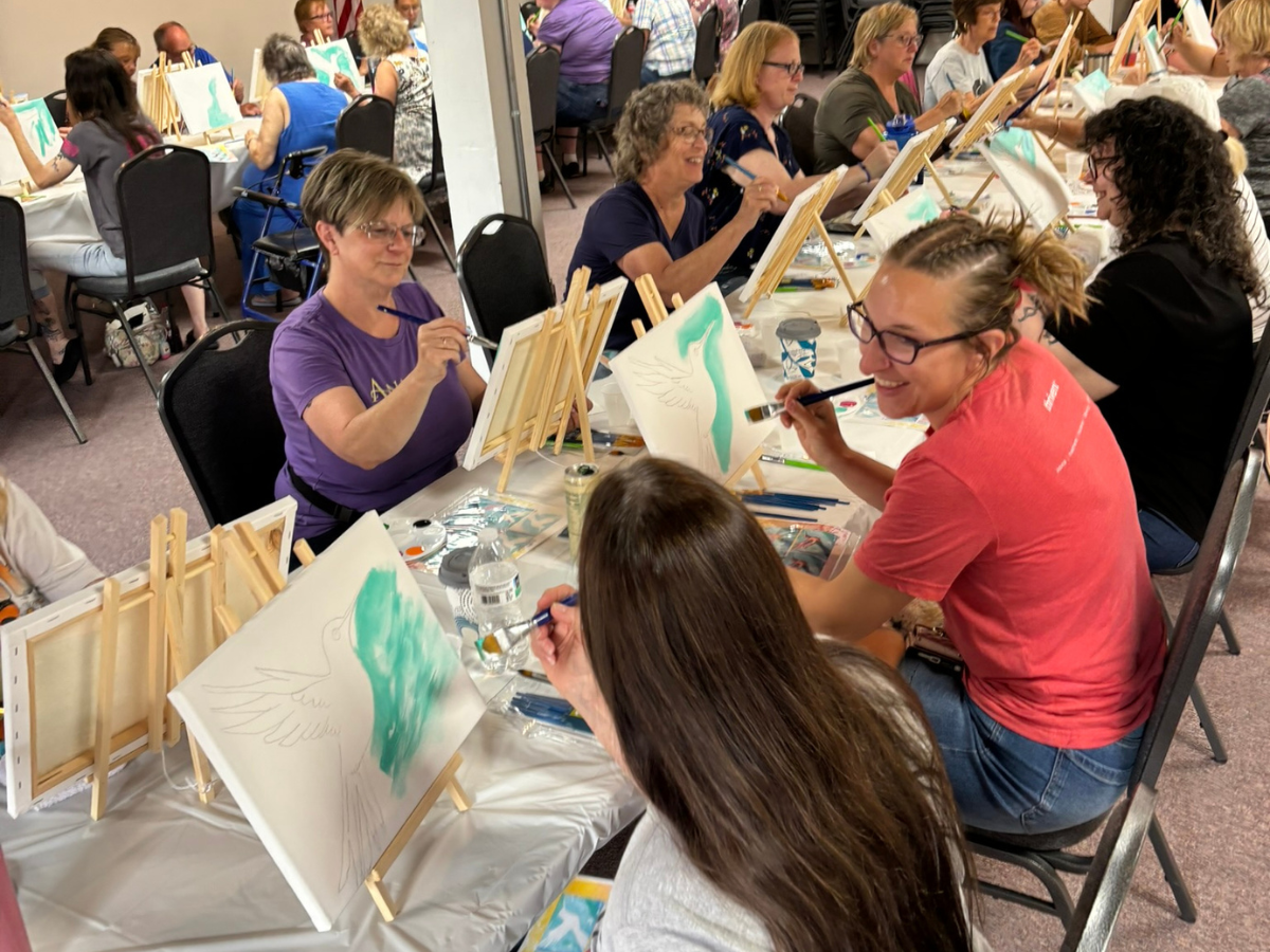 A group of people seated at long tables, painting on canvas easels in a large indoor event space. The foreground shows two women painting, with more participants and artwork visible in the background.