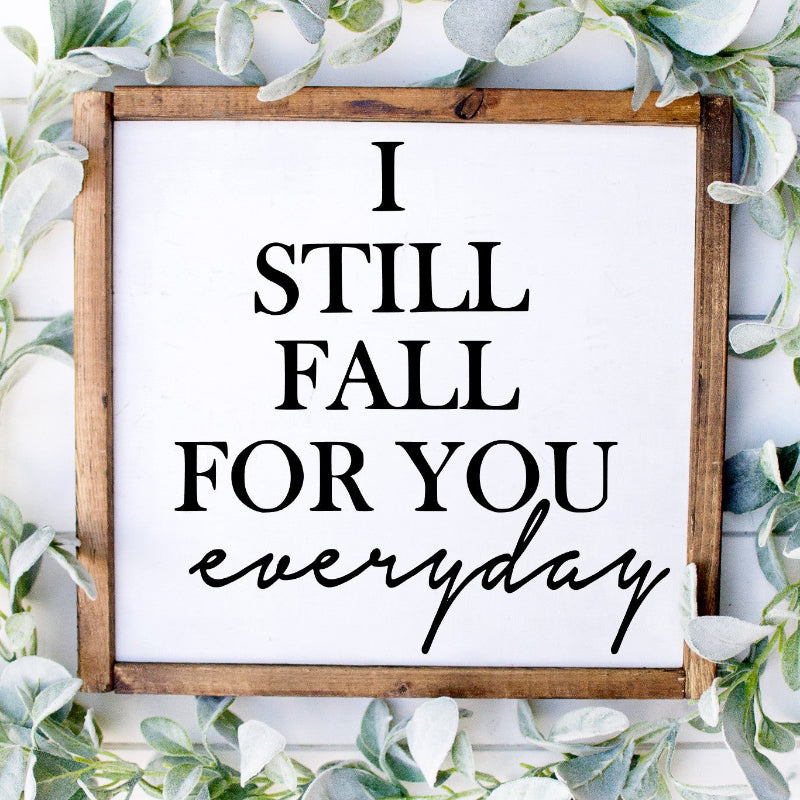 Wooden sign with 'I STILL FALL FOR YOU EVERYDAY' text surrounded by greenery on a white background