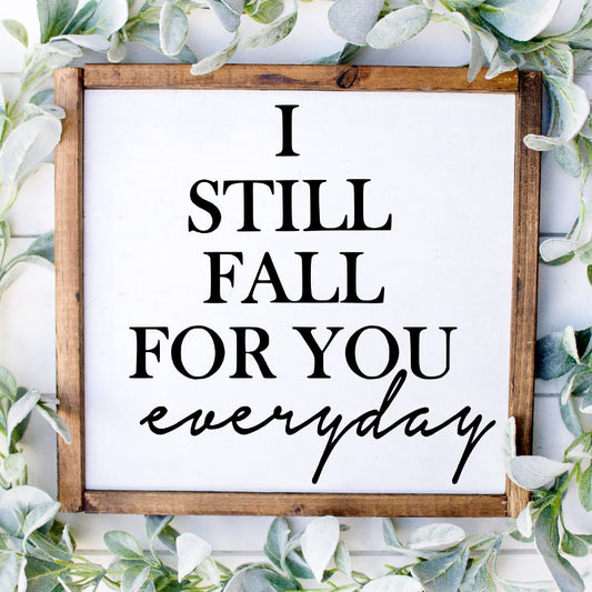 Wooden sign with 'I STILL FALL FOR YOU EVERYDAY' text surrounded by greenery on a white background