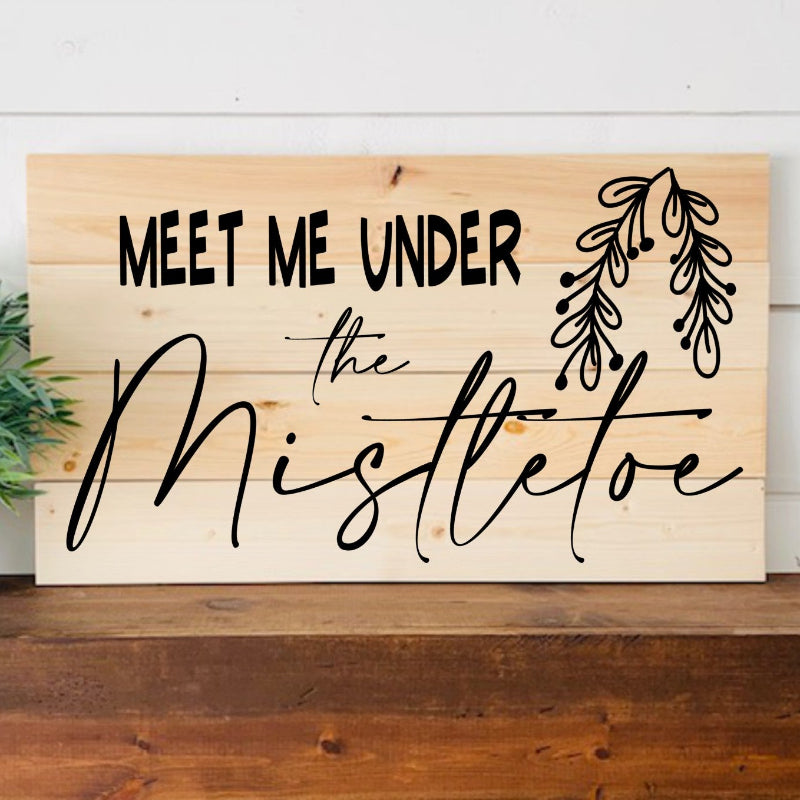 Wooden sign with 'Meet Me Under the Mistletoe' text on a wooden surface with a plant in the background.