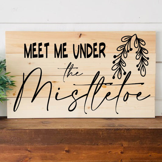 Wooden sign with 'Meet Me Under the Mistletoe' text on a wooden surface with a plant in the background.