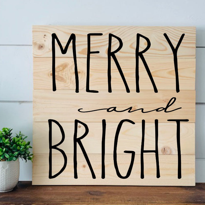 Wooden sign with 'Merry and Bright' text on a wooden surface with a plant in the background.