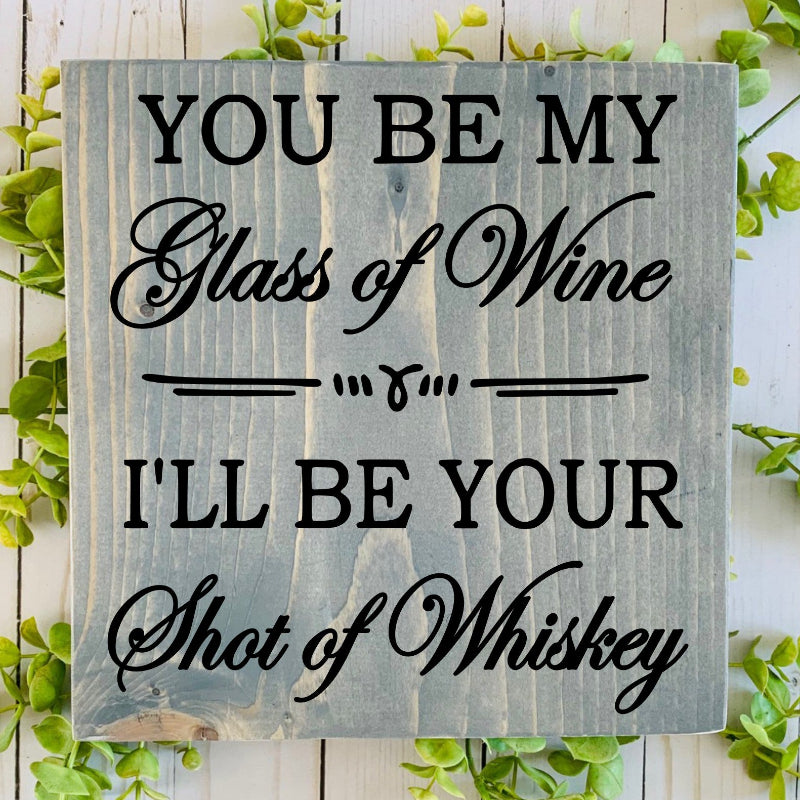 Wooden sign with text 'You be my glass of wine, I'll be your shot of whiskey' on a wooden background with greenery.